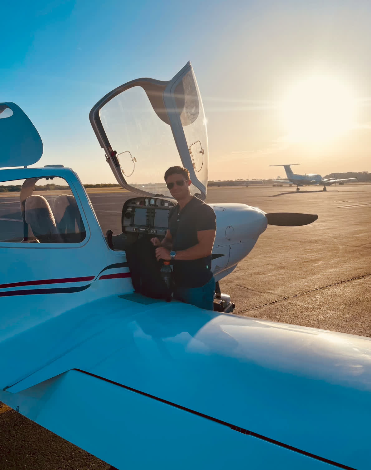 Aaron Howard standing beside a light aircraft on the tarmac at sunset, wearing a headset